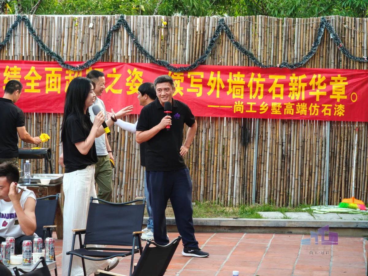 福建尚工乡墅装饰工程有限公司团队活动，员工在“尚工乡墅高端仿石漆”和“外墙仿石新华章”业绩横幅前登台发言。